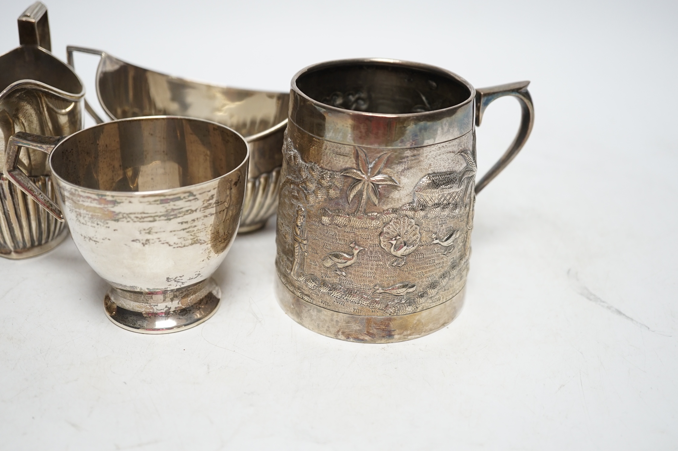 Two early 20th century demi-fluted silver cream jugs and a similar sugar bowl, an Irish silver tea cup and an Indian? white metal mug, gross weight 16.7oz. Fair condition.